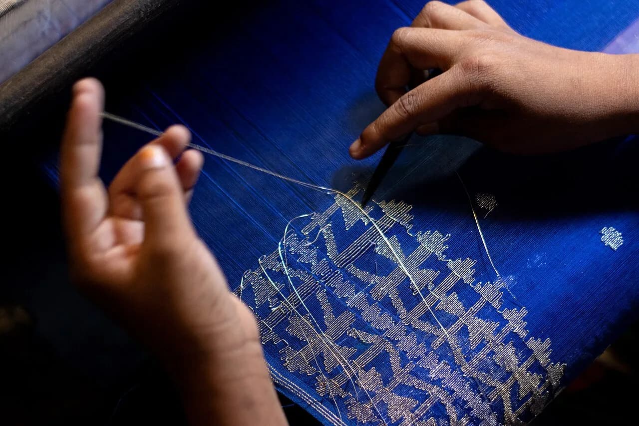 Mani al lavoro su un tessuto Jamdani blu con motivi chiari.